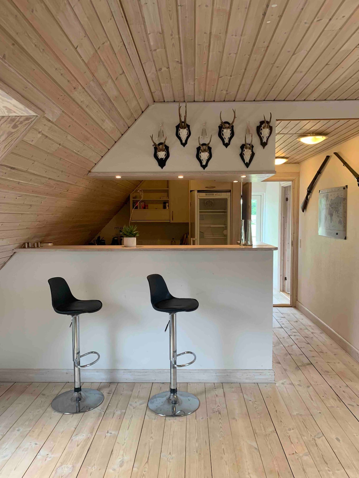 A bar area with two modern black stools stands in front of a minimalist counter. Decorative wall mounts featuring antlers are displayed above the counter, contributing to the rustic ambiance. Natural light filters in, highlighting the wooden ceiling and light-colored floor.
