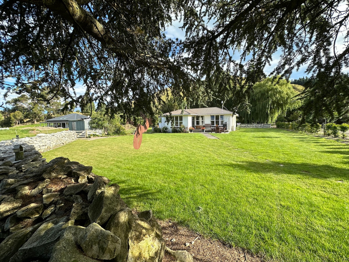 The cottage is framed by a large, grassy lawn bordered by established trees and a stone wall. The single-story building, with several windows, is set amidst a peaceful landscape of hills and fields, inviting relaxation and exploration.