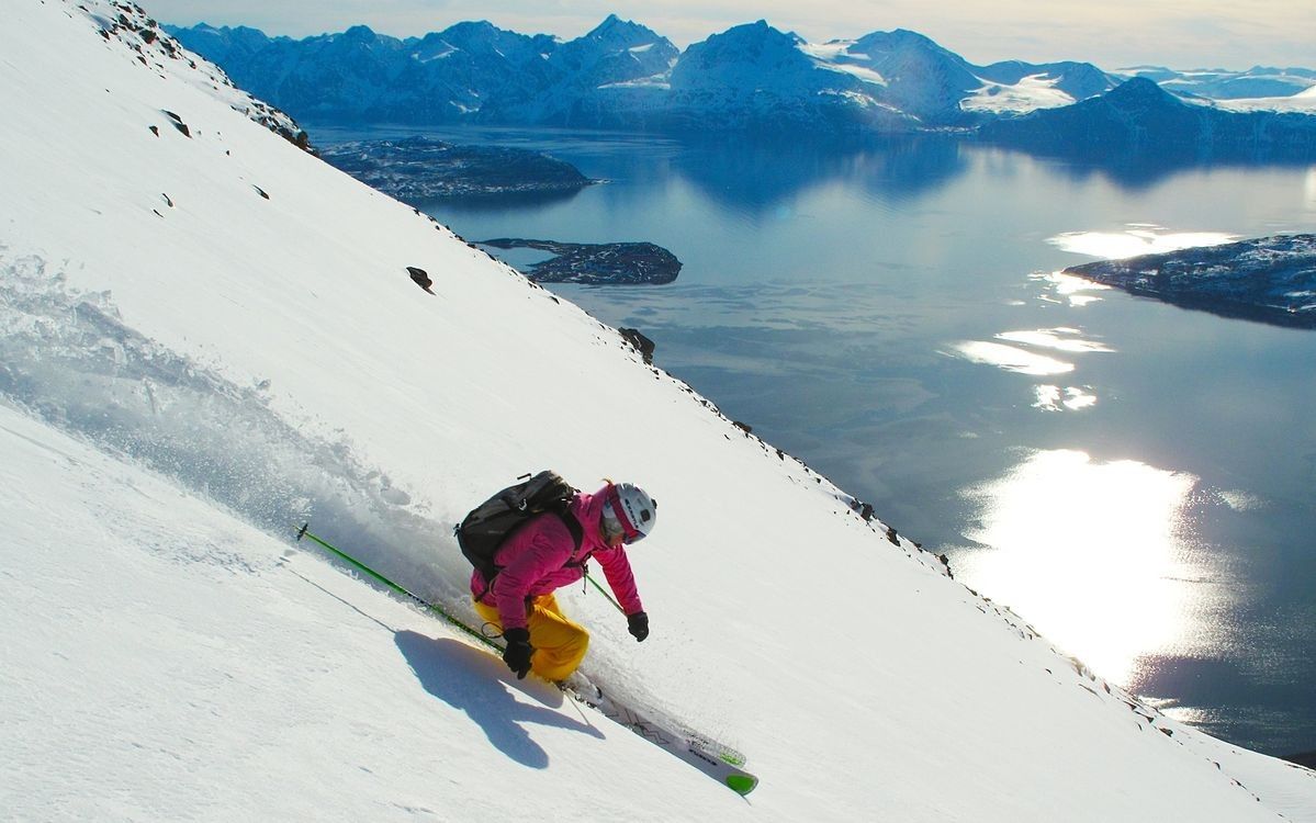 A skier descends a snowy slope, with bright pink and yellow clothing contrasting against the white landscape. The expansive view reveals a serene body of water reflecting sunlight, surrounded by majestic mountains in the distance.