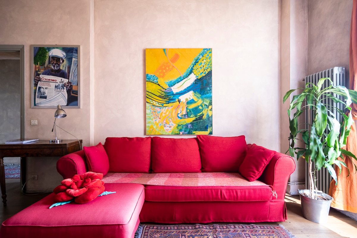 A red sofa and ottoman are positioned in a sunlit corner, complemented by a vibrant artwork above. A wooden table and lamp are nearby, with a potted plant to the side, adding a touch of greenery to the space.
