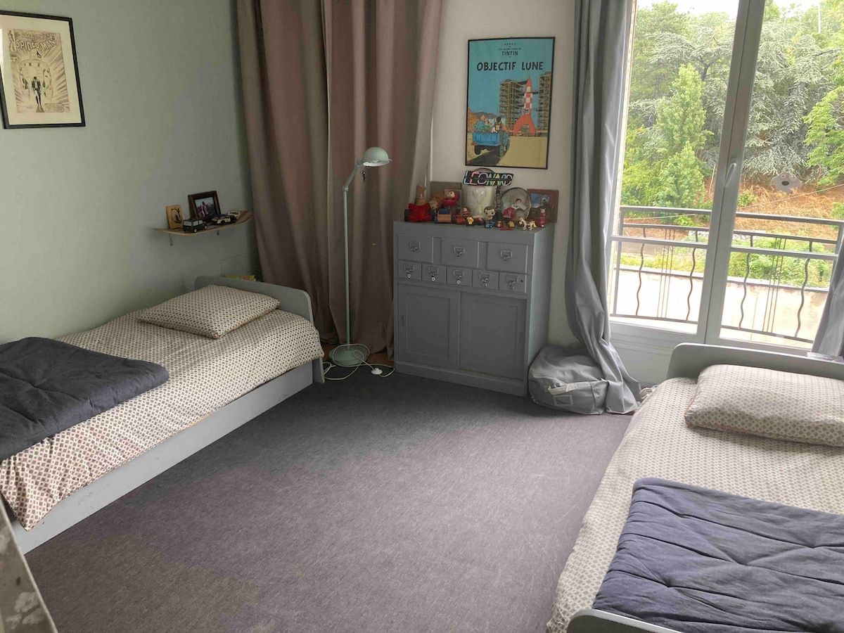A modern bedroom features two twin beds positioned on a soft carpet. A tall dresser stands against one wall, while a reading lamp provides light. Large windows are covered by flowing curtains, revealing views of greenery outside.
