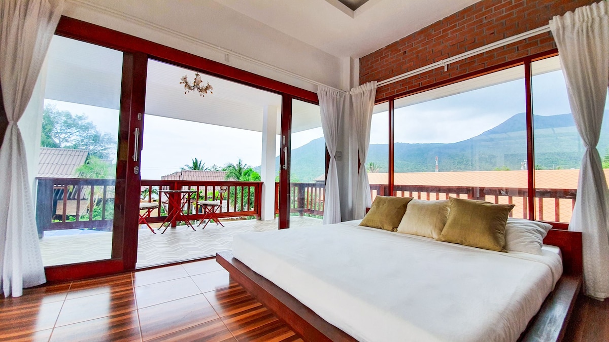 A spacious sleeping area features a low-profile bed with soft bedding and decorative pillows. Large glass doors open to a private balcony, offering mountain views and access to outdoor seating. Natural light fills the room, highlighting the warm wooden floors and exposed brick walls.