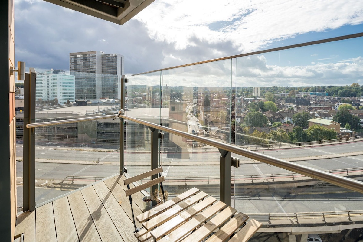 A balcony is seen with a wooden slatted table and seating offering an unobstructed view of the cityscape. Glass railings reflect natural light, and cloudy skies are visible above, creating a serene atmosphere.