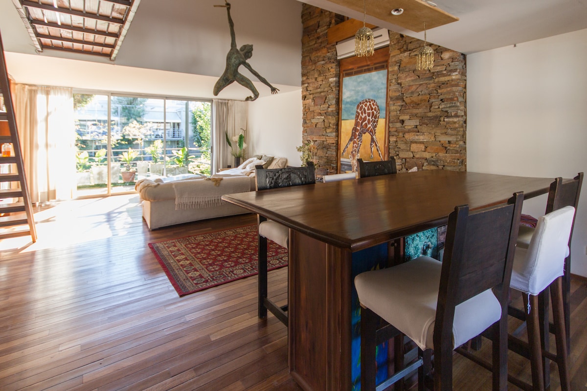 A bright living area features a spacious seating arrangement complemented by large glass doors, which provide views of the outdoor space. A wooden dining table with several chairs is positioned in the foreground. A decorative wall art piece and a nature-inspired sculpture enhance the room's character.