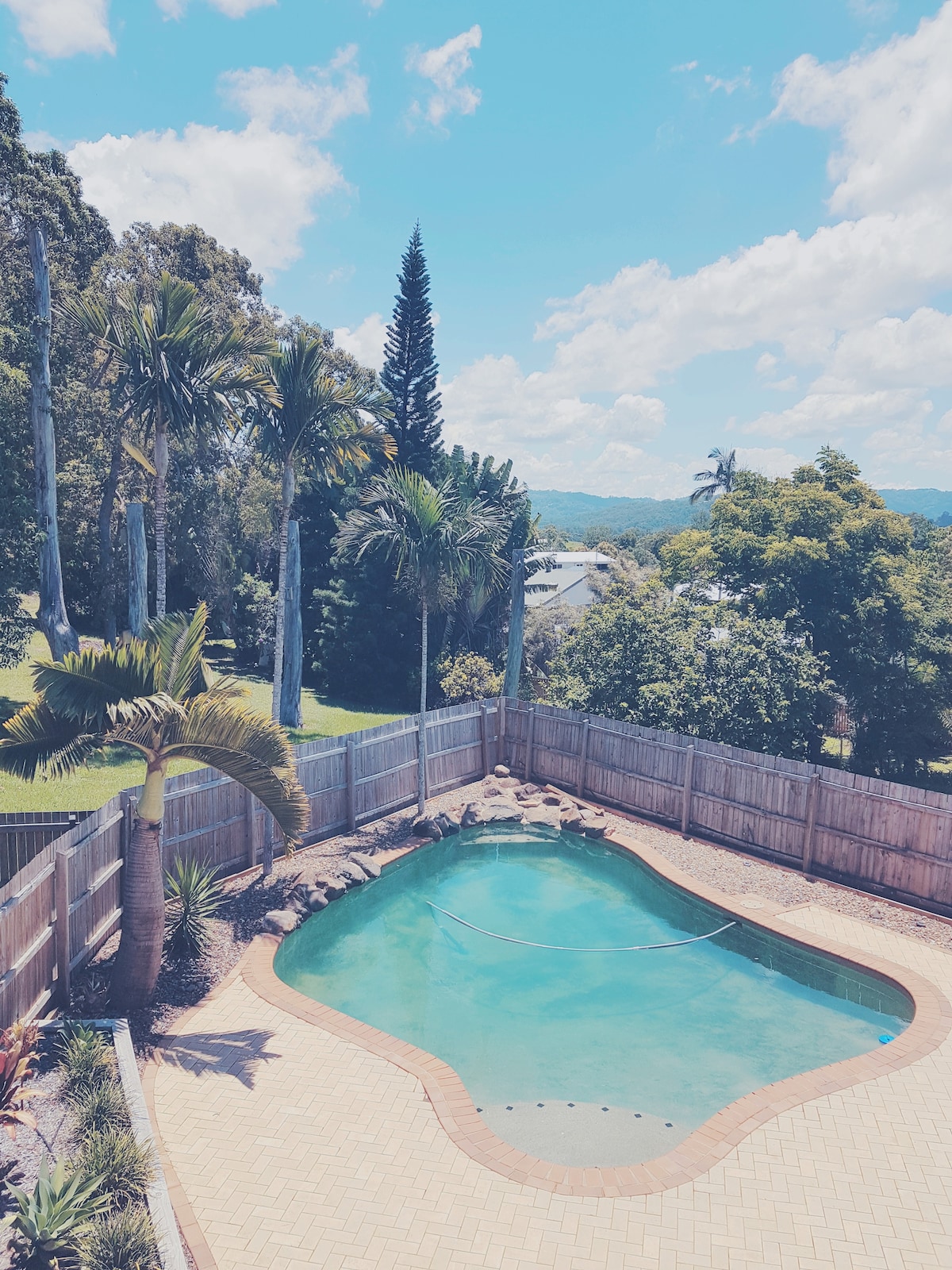 A clear view of a private pool is visible, surrounded by tropical landscaping and trees. The pool is shaped organically, with a light blue hue reflecting the sky. A wooden fence borders the area, providing a sense of seclusion against the backdrop of lush greenery and distant hills.