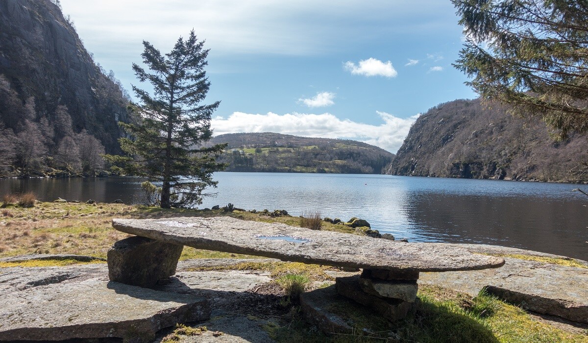 A tranquil lakeside scene features a large flat stone table resting on natural stone supports, surrounded by grassy areas. In the background, a serene lake reflects gentle hills, under a sky with scattered clouds and patches of blue. Trees line the shore, enhancing the peaceful landscape.