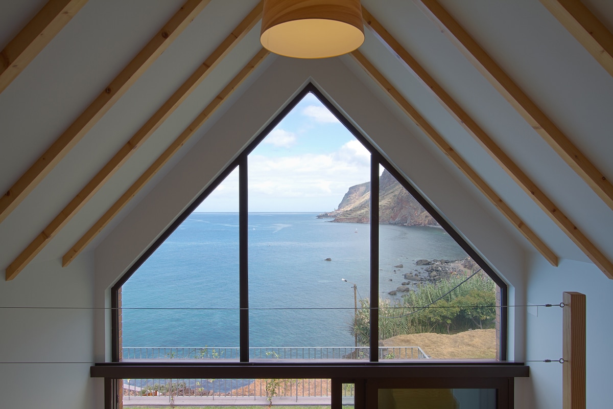 A large triangular window showcases expansive views of the coastline, with gentle waves lapping against the shore. Natural light fills the space, highlighting the wooden beams of the ceiling and offering a serene connection to the outside landscape.