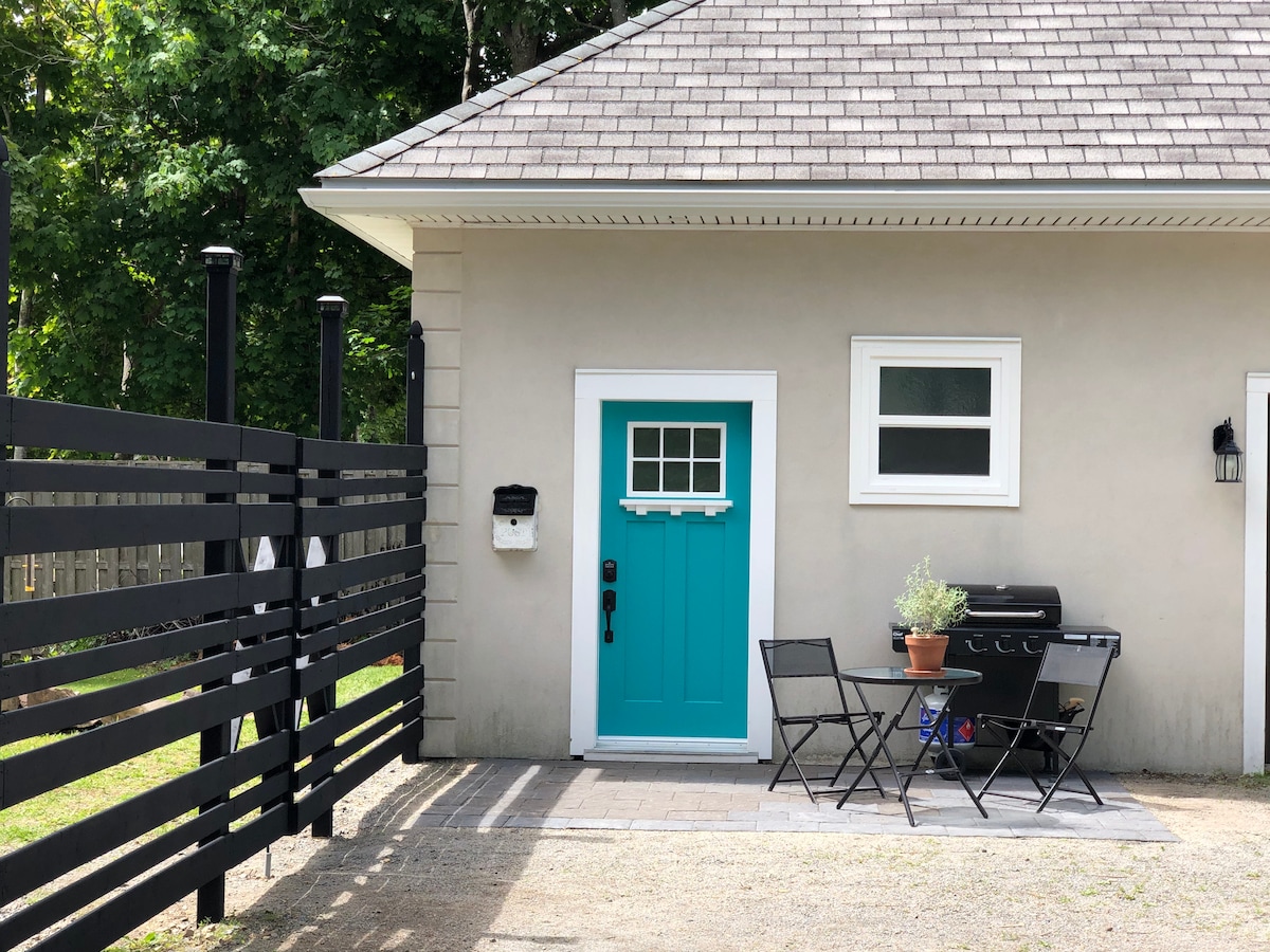 A private entrance is highlighted by a vibrant turquoise door, featuring a small window above the door handle. An outdoor patio area includes a bistro table with two chairs and a BBQ grill, surrounded by a modern black fence that enhances privacy.