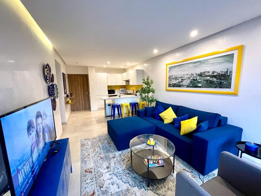 A modern living area features a plush blue sectional couch adorned with bright yellow cushions. A glass coffee table rests on a patterned area rug. A large television is positioned in front, and the kitchen is visible in the background with sleek cabinetry and island seating.