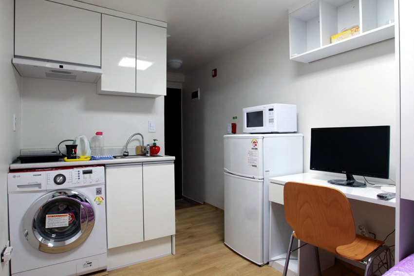 A modern kitchen area features sleek cabinetry and essential appliances, including a washing machine, refrigerator, and microwave. A small desk with a monitor is positioned beside the kitchen. Natural light brightens the space, enhancing the functional design.