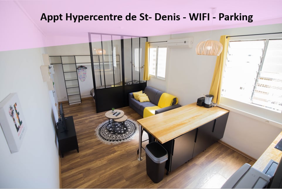 An open living area features a seating space with a dark sofa accented by yellow cushions. A light wooden table is positioned in the foreground, alongside a black and yellow kitchen setup. Large windows allow natural light to brighten the room, enhancing the modern design.