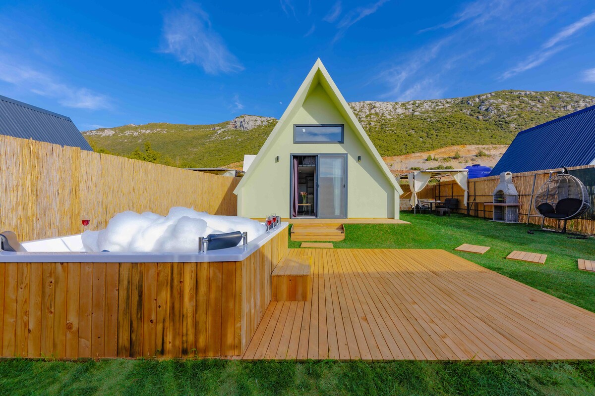 A modern A-frame structure is set against a backdrop of rolling hills and a clear blue sky. Wooden decking surrounds a hot tub, while a grassy area leads to tall bamboo fencing. Large glass doors provide access to the inviting indoor space.