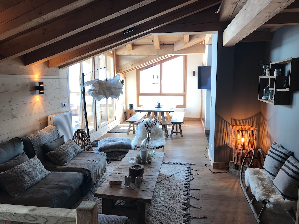 The spacious living area features a comfortable sectional sofa and a wooden coffee table, blending with natural wooden elements. Large windows allow ample natural light, and a dining table is visible in the background. Soft textiles and decorative items add a cozy touch to the space.