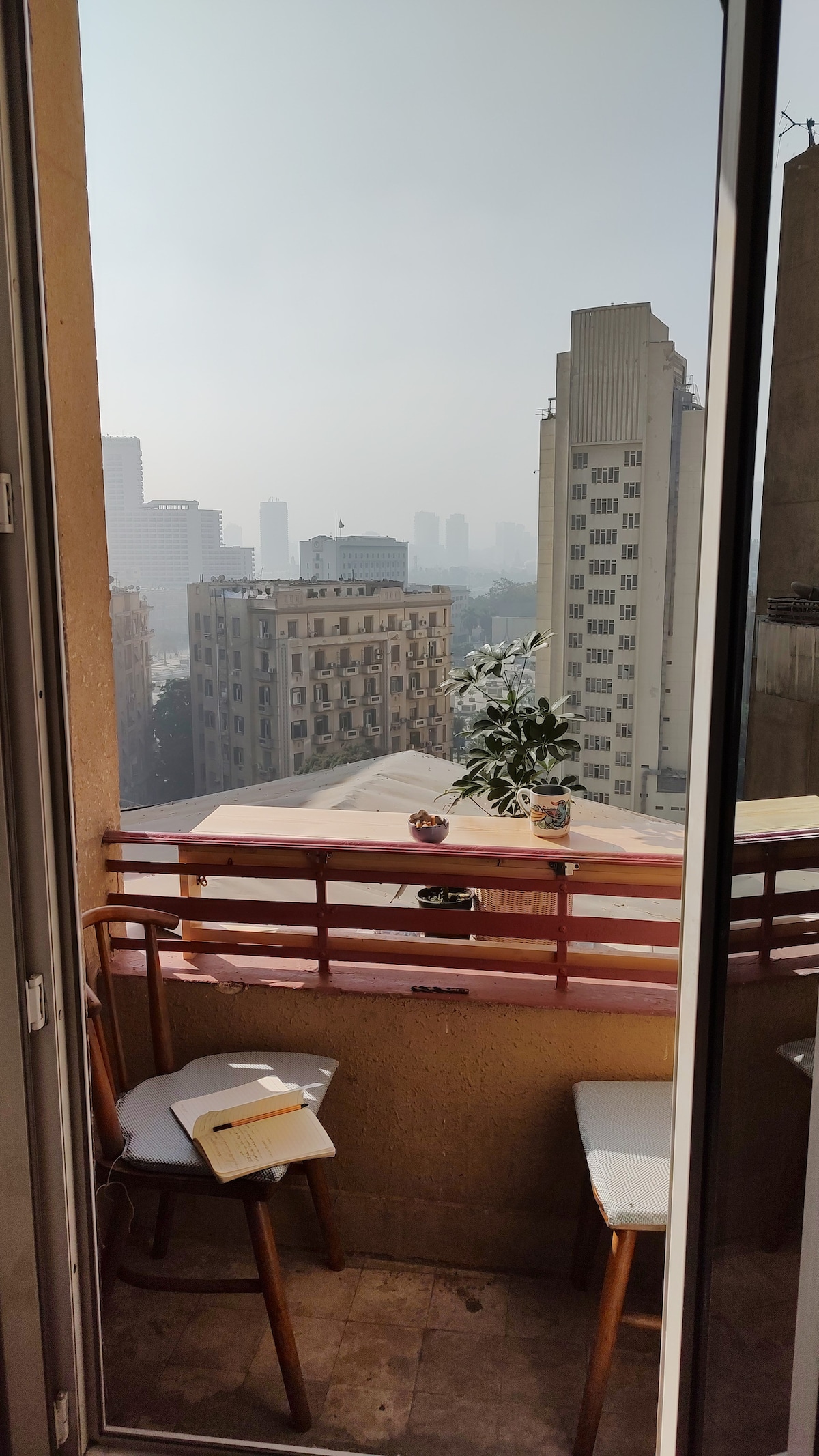 A small balcony is visible, featuring two wooden chairs and a small table with a potted plant. The view encompasses the skyline of Downtown Cairo, showcasing a mix of modern buildings and architecture, enveloped in a soft haze.