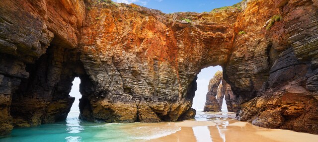The image features a scenic beach landscape with distinct rock formations creating natural arches. Soft sand stretches along the shoreline, while turquoise waves gently lap at the base of the rocks. Bright blue skies complement the vibrant colors of the rocky cliffs.