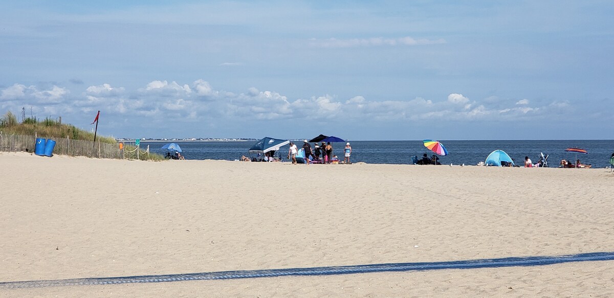 A wide expanse of sandy beach stretches toward the water's edge, where a gentle sea meets a blue sky dotted with clouds. Beachgoers are seen lounging under various colorful umbrellas, while distant small tents provide shade. A few people can be spotted enjoying the shore.