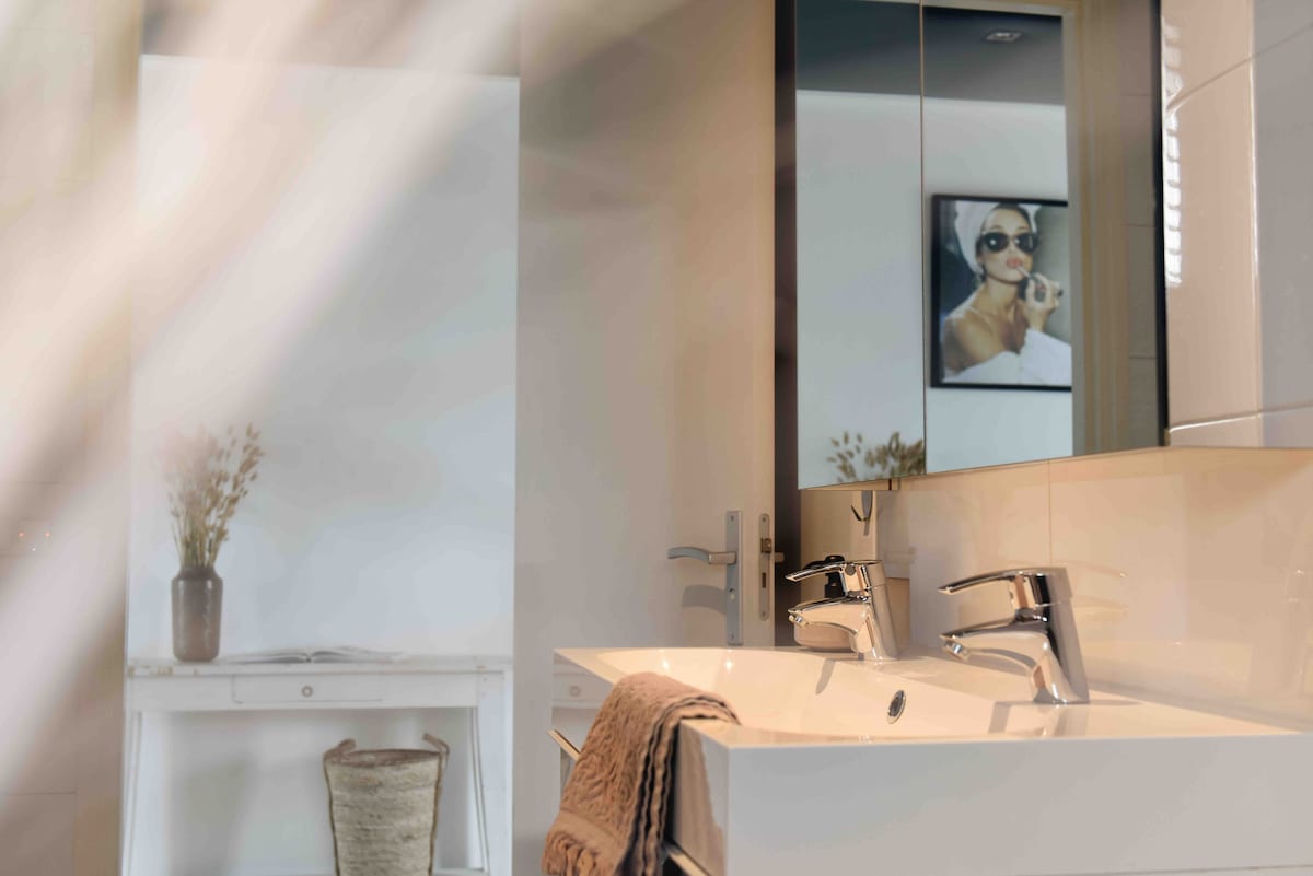 The bathroom features a double vanity with sleek chrome faucets and a minimalistic design. A mirror reflects an artistic image on the wall, while a small vase with dried flowers adds a touch of decor. Soft textiles are draped over the countertop.