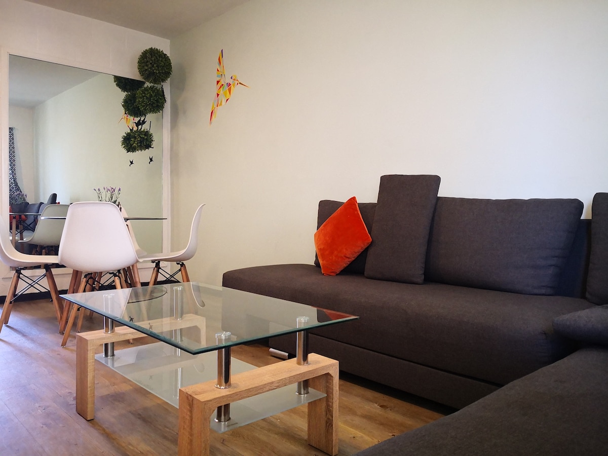 A modern living area features a spacious dark gray sofa with an orange accent pillow. A glass coffee table sits on a wooden floor, reflecting natural light. In the background, a dining table with white chairs is visible, along with decor on the walls.