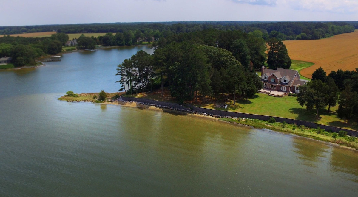 An aerial view captures the large waterfront estate surrounded by water on two sides. Lush greenery borders the property, which features a spacious lawn leading up to the home. The serene waters reflect the sky, while cultivated fields extend into the distance.