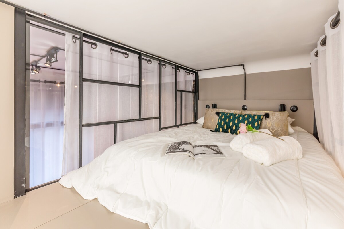 An intimate sleeping area is positioned on a mezzanine, featuring a spacious bed adorned with soft linens and decorative pillows. Large windows framed by sheer curtains allow natural light to enter, creating a bright and airy atmosphere.
