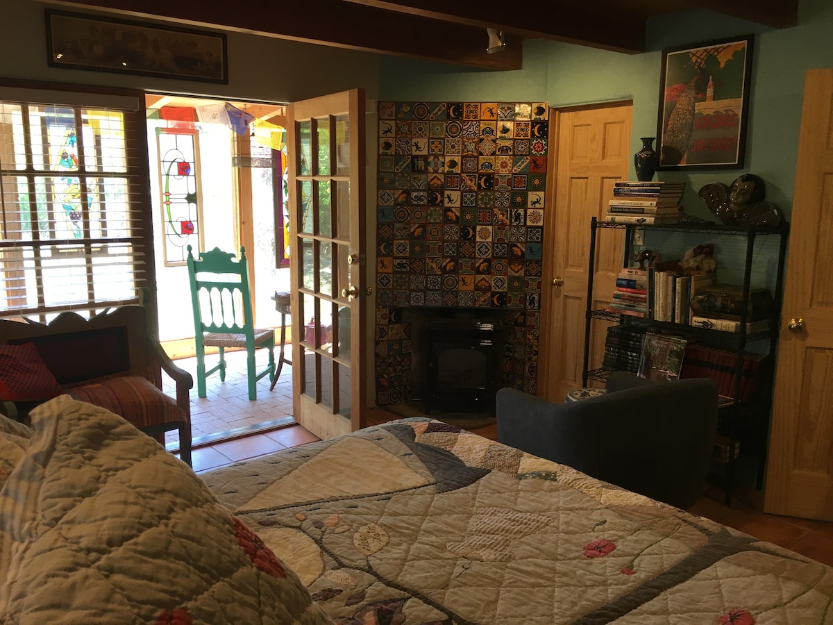 A cozy sleeping area is highlighted by a spacious bed with patterned bedding. Colorful tiles adorn a feature wall, complemented by a bookshelf filled with books. A gas fireplace adds warmth, while a separate area showcases a sunlit enclosed porch through large glass doors.