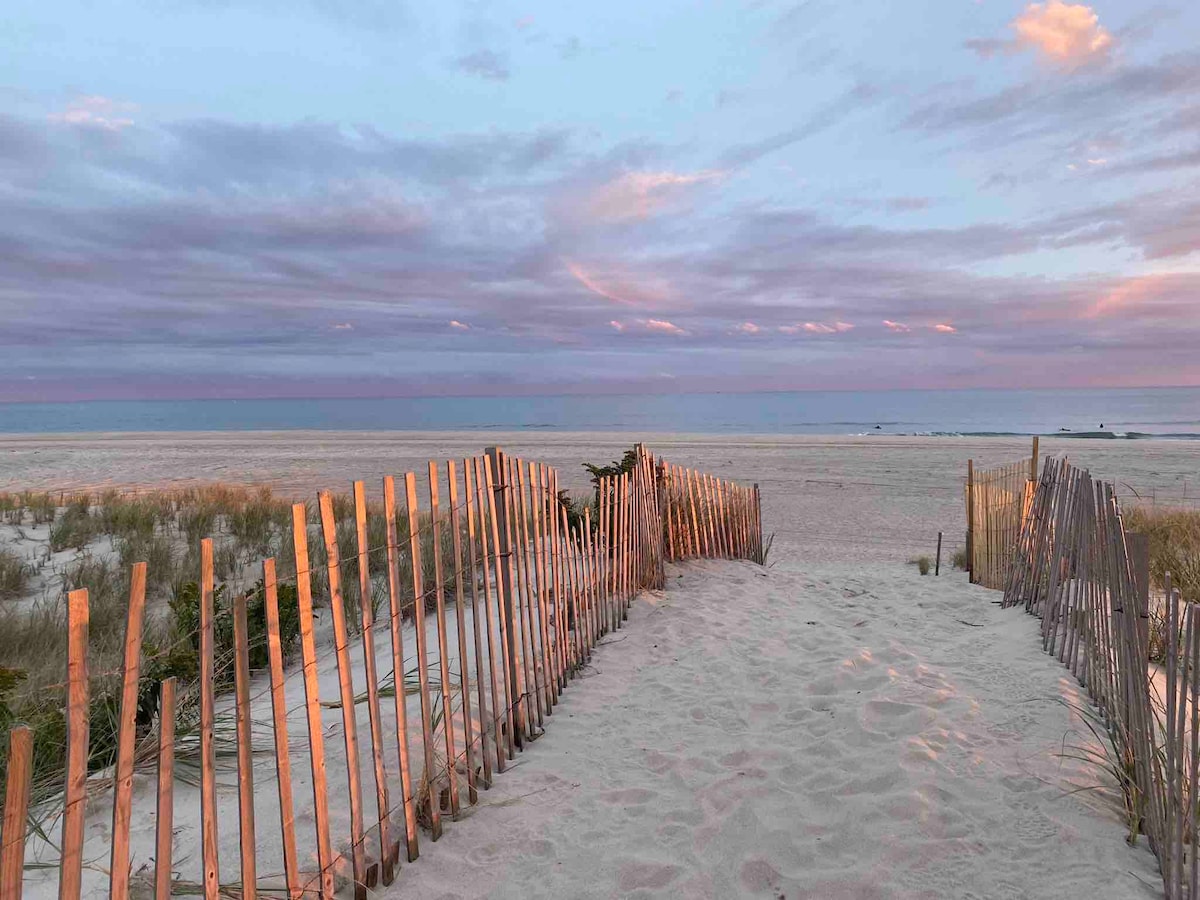 A wooden fence made of slender posts lines a sandy path leading to the beach, framed by tranquil grasses. Soft pastel hues fill the sky at dusk, transitioning beautifully above the calm ocean waves in the distance.