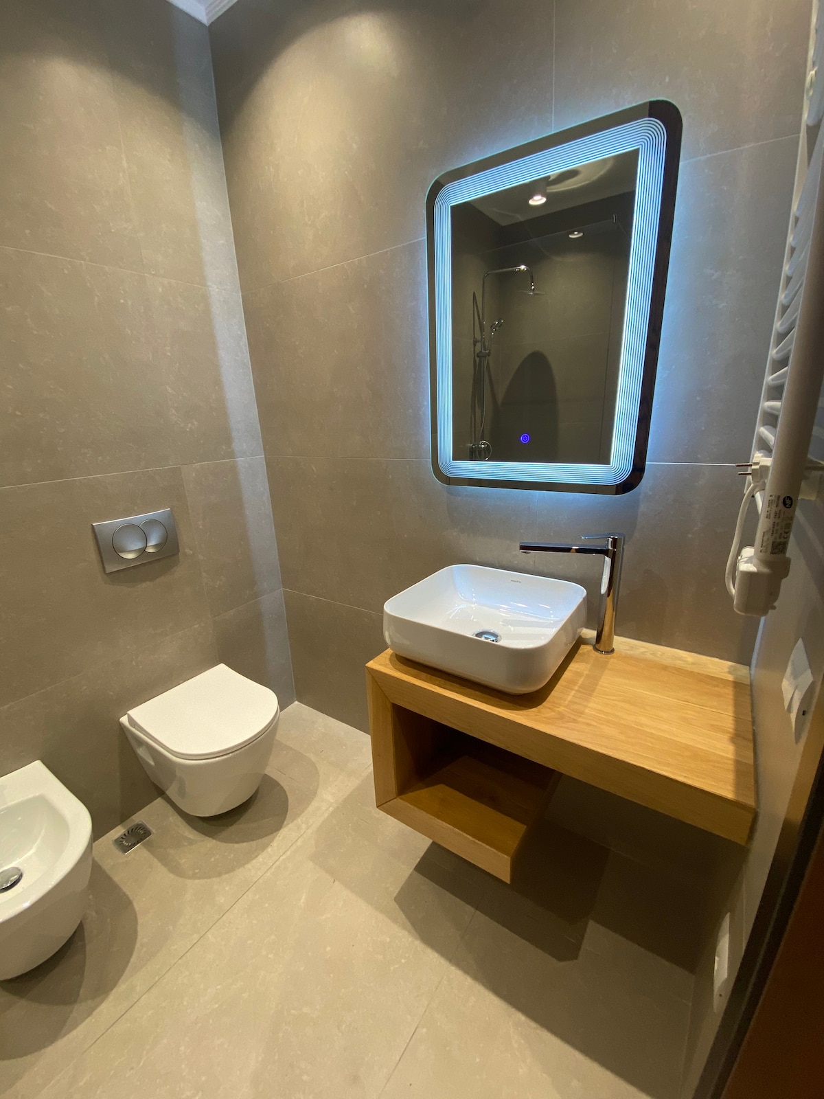 A modern bathroom is presented with neutral-toned tiles. A stylish, illuminated mirror hangs above a minimalist sink on a wooden vanity. A compact toilet and a bidet are positioned beside each other, contributing to the clean and efficient layout of the space.