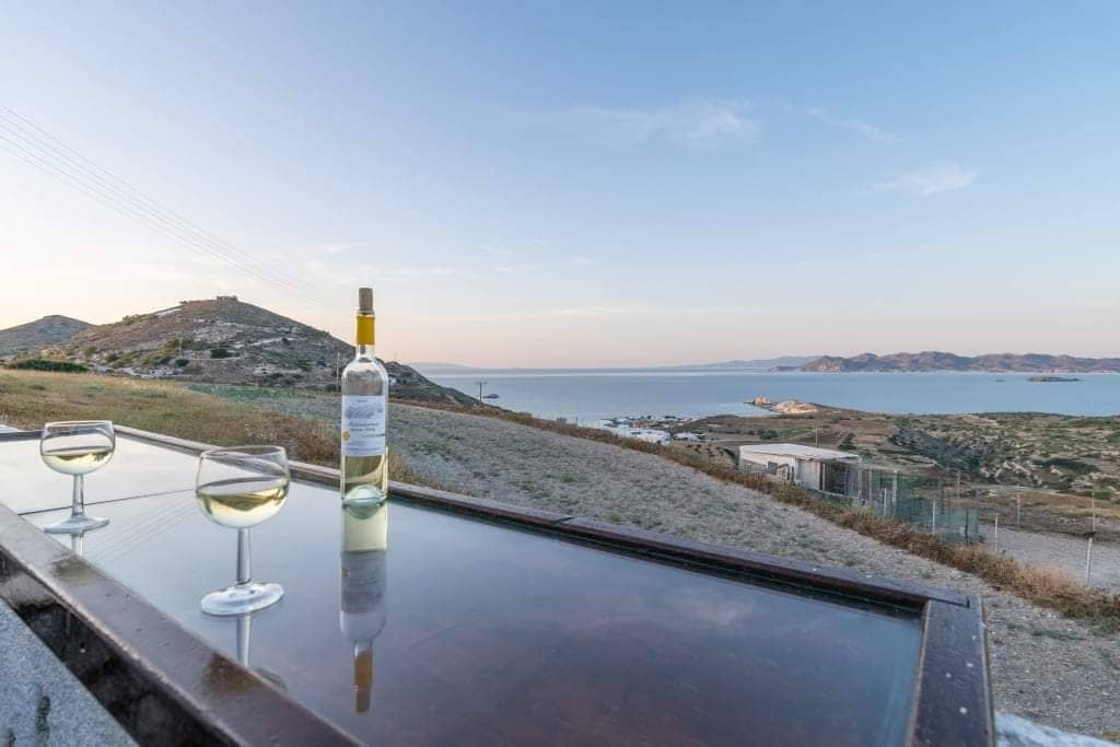 A scenic view features a bottle of white wine placed on a polished table, accompanied by two glasses filled with wine. The landscape includes rolling hills and the blue waters extending to the horizon under a soft sky.