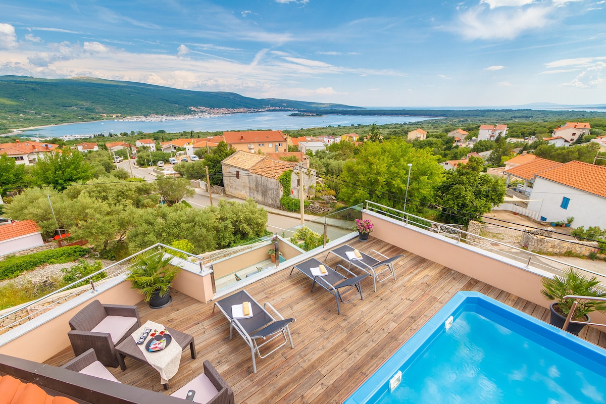 A spacious roof terrace features a swimming pool and several lounge chairs arranged around a small table. Lush greenery and views of the village and coastline are prominent in the background, with a serene sky above and the sea extending into the distance.