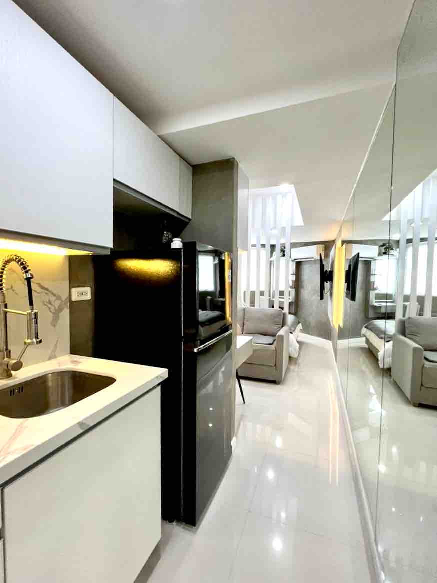 A modern kitchen area is visible, featuring a sink and a black refrigerator. Smooth, shiny countertops reflect light, while a spacious living area can be seen in the background. Mirrored walls enhance the sense of space and brightness throughout the room.
