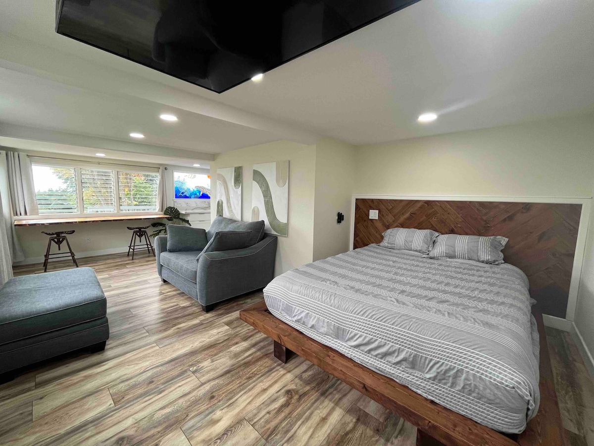 A spacious interior features a queen bed with a patterned headboard against one wall. Seating areas include a gray couch and a chaise lounge. Large windows offer natural light, with a view of the outdoors, while a Smart TV is mounted across the room.
