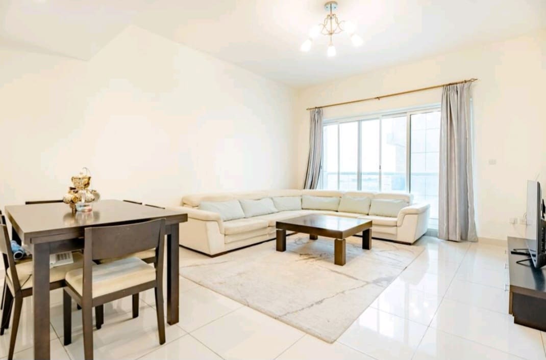 A light-filled living area features a spacious sectional sofa and a central coffee table set on a soft area rug. A dining table for four is positioned nearby. Large windows allow natural light to brighten the space, complemented by neutral wall tones.