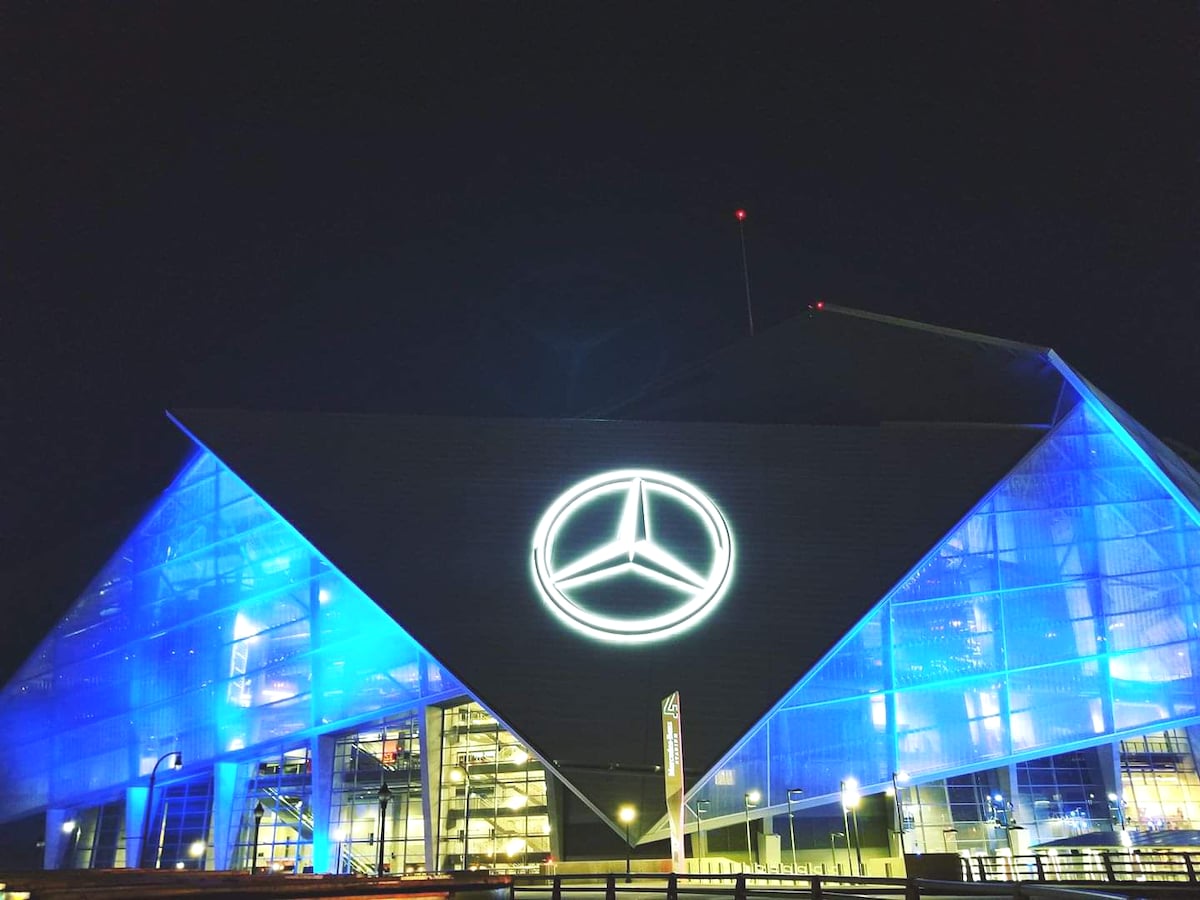 The exterior of Mercedes Benz Stadium is illuminated at night with vibrant blue lighting, highlighting its modern architectural design. The iconic Mercedes logo is prominently displayed, creating a focal point on the building's facade.