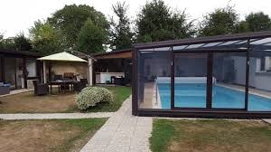 A garden area is featured with a swimming pool enclosed by glass walls. A shaded seating area under a green canopy is visible, alongside outdoor furniture positioned on a stone path leading to the pool. Lush trees provide natural privacy around the space.