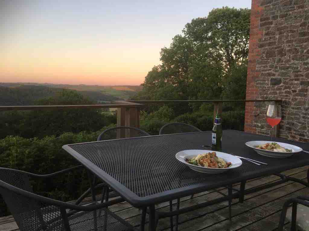 A dining table is set outdoors, surrounded by metal chairs. Two plates of food and a glass of drink are positioned on the table, with a scenic sunset view in the background, displaying rolling hills and a colorful sky.