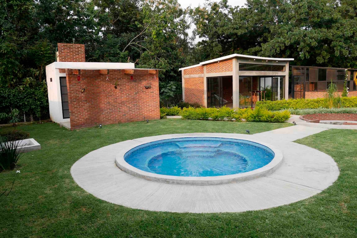 A private outdoor space features a circular swimming pool surrounded by a neatly manicured lawn and lush greenery. Two modern brick structures are visible in the background, with large windows allowing for natural light. The area is designed for relaxation and enjoyment.