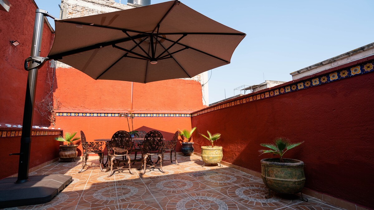 A private terrace is highlighted, featuring a large umbrella over a dining table with ornate metal chairs. Potted plants add greenery against vibrant red walls, with intricate tiling along the edge. Sunlight bathes the space, creating a warm and inviting outdoor setting.