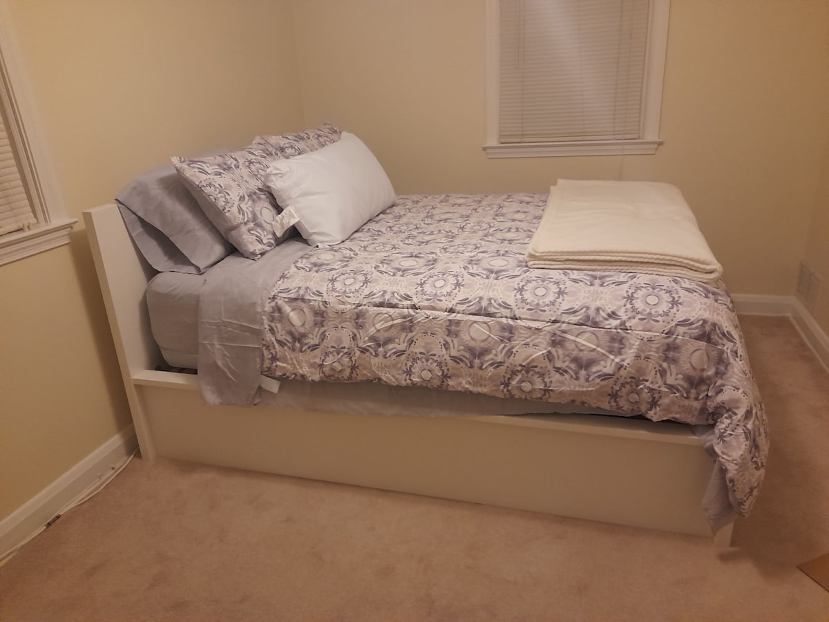 A neatly arranged sleeping area features a raised bed with a patterned comforter and matching pillows. A soft blanket is placed at the foot of the bed. Warm overhead lighting enhances the light-colored walls and soft carpeting.