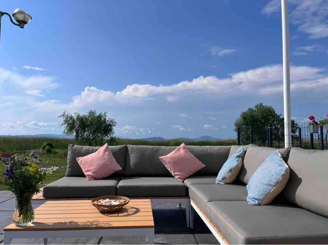 A spacious outdoor seating area is presented, featuring a sectional sofa adorned with pastel-colored cushions. A wooden table is positioned in front, accompanied by a decorative bowl. The backdrop features a clear blue sky and distant greenery, creating a peaceful environment.