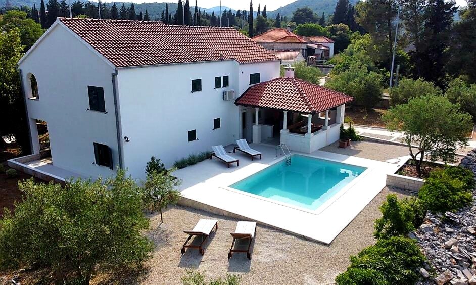 A spacious two-story house features a traditional tiled roof and green window shutters. A private swimming pool is surrounded by a sun terrace with lounge chairs. Nearby, a covered terrace with a wooden structure provides shaded seating, complemented by the lush greenery of the surrounding garden.