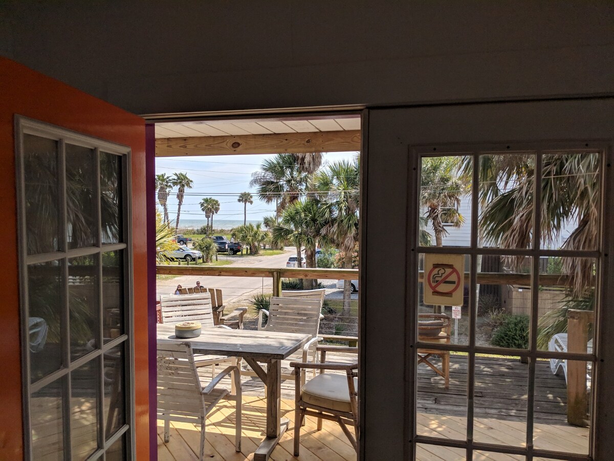 An open doorway frames a spacious deck with wooden furniture overlooking the ocean. Palms are visible in the background, along with a glimpse of the beach. Natural light flows into the interior, enhancing the warm, weathered tones of the space.