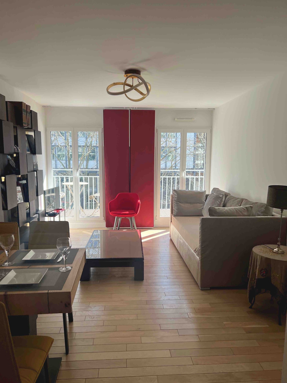 A bright living area is presented, showcasing a spacious grey sofa and a red accent chair. A wooden dining table is set with dishes, and a black lamp stands on a side table. Large windows allow natural light to fill the room, highlighting the wooden flooring.