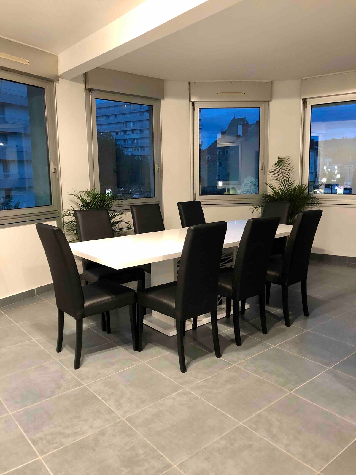 A bright dining area features a large white table surrounded by six black chairs. Large windows allow soft light to illuminate the space, offering views of the surrounding environment. Potted plants add a touch of greenery, enhancing the atmosphere.