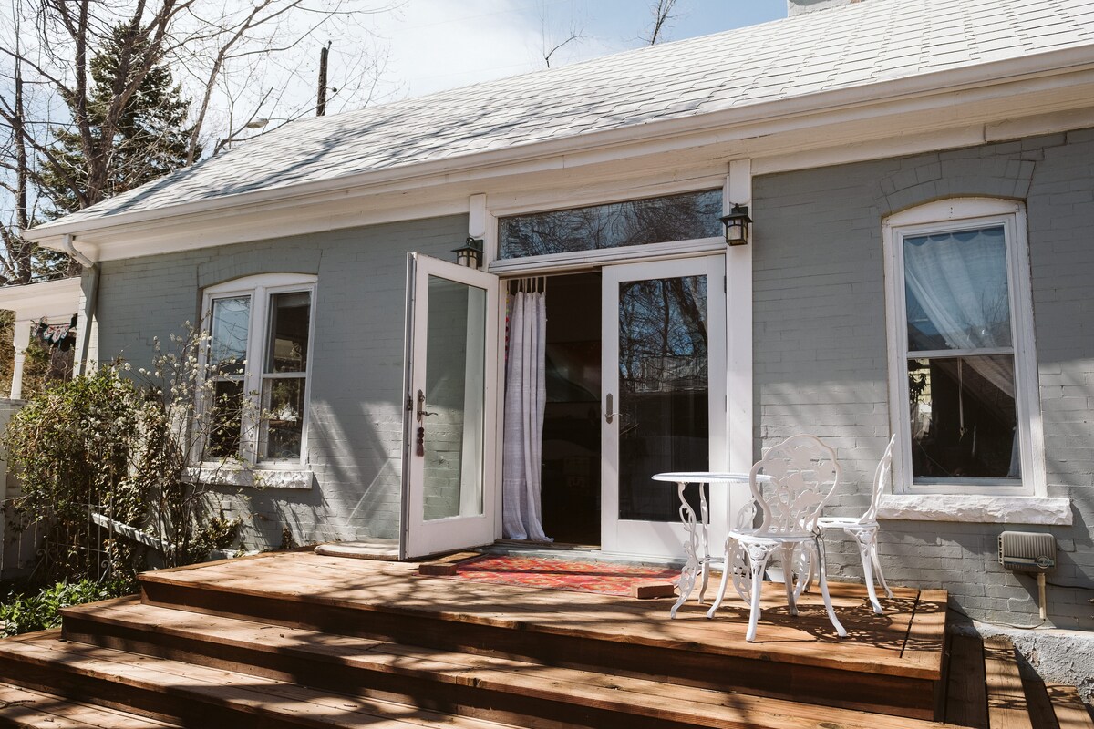 An inviting entrance welcomes guests with a classic wooden deck leading to double glass doors. A small white table and chairs rest on the porch, surrounded by blossoming greenery. Soft light filters through sheer curtains, enhancing the charm of the bungalow's façade.