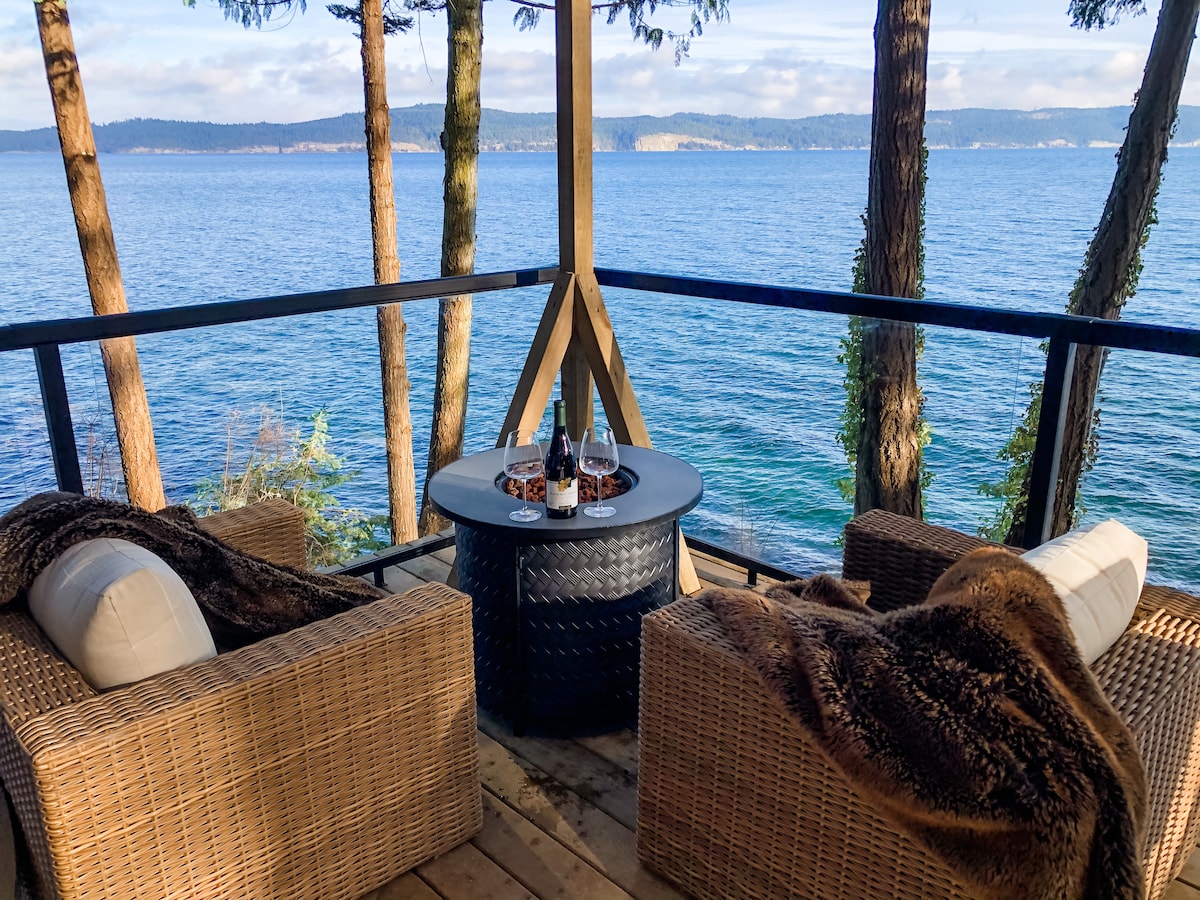 An outdoor seating area is featured with two woven chairs positioned around a circular fire pit. Plush throws are draped over the chairs, and the backdrop consists of tall trees with scenic ocean views beyond the railing.
