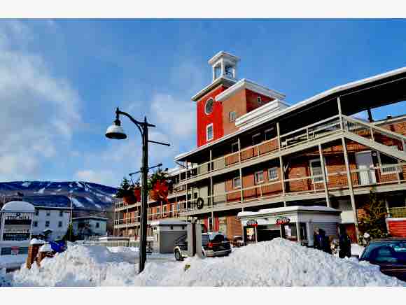 The newly remodeled condo is situated in a historic Wool Mill building, characterized by a red brick facade. The building features a distinctive clock tower and multiple levels, with balconies visible on the second floor. Snow-covered ground enhances the winter scene.