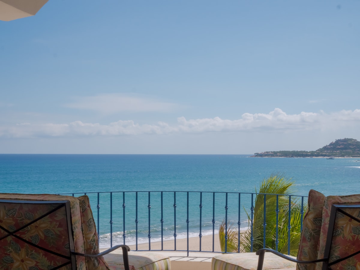 A panoramic ocean view is captured from the balcony, showcasing vibrant blue waters meeting a clear sky. The railing provides a sense of openness, while two patterned chairs face the sea, creating a quiet spot for relaxation.