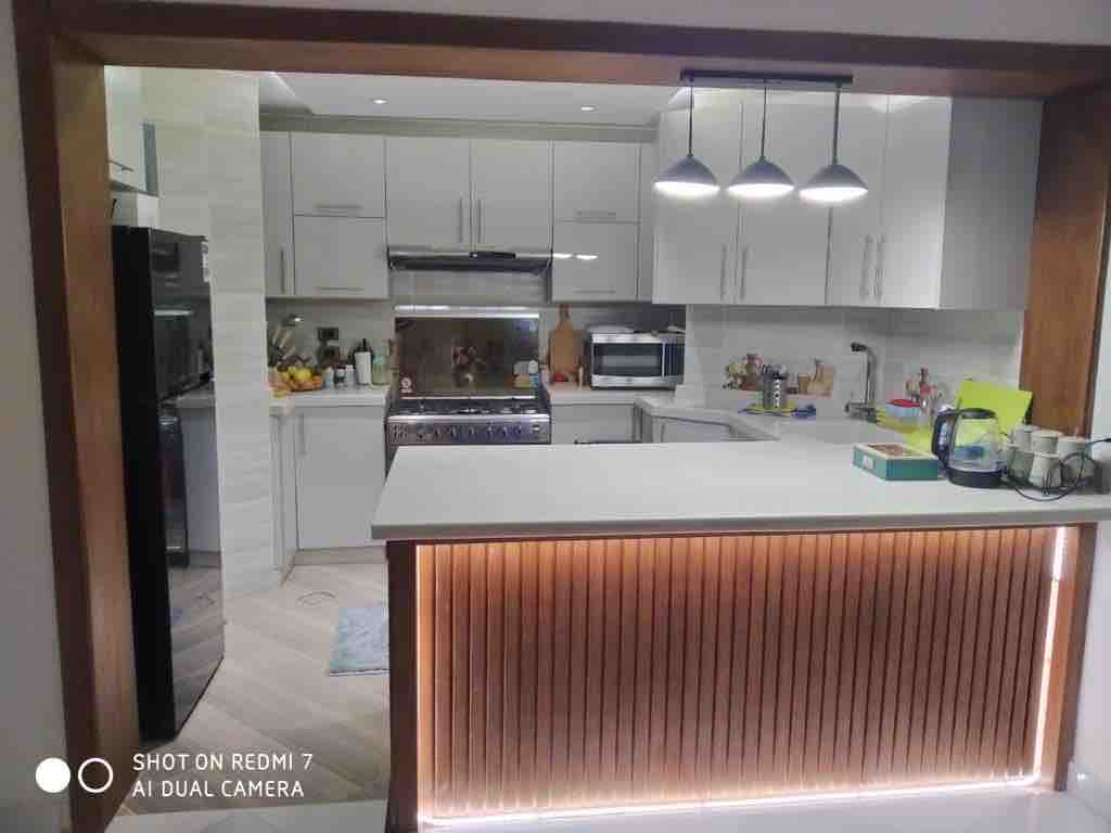 A modern kitchen features sleek white cabinetry and a spacious central island with warm wood accents. Stainless steel appliances, including a refrigerator and oven, are visible, along with various items arranged on the countertops. Under-cabinet lighting highlights the clean lines and contemporary design.