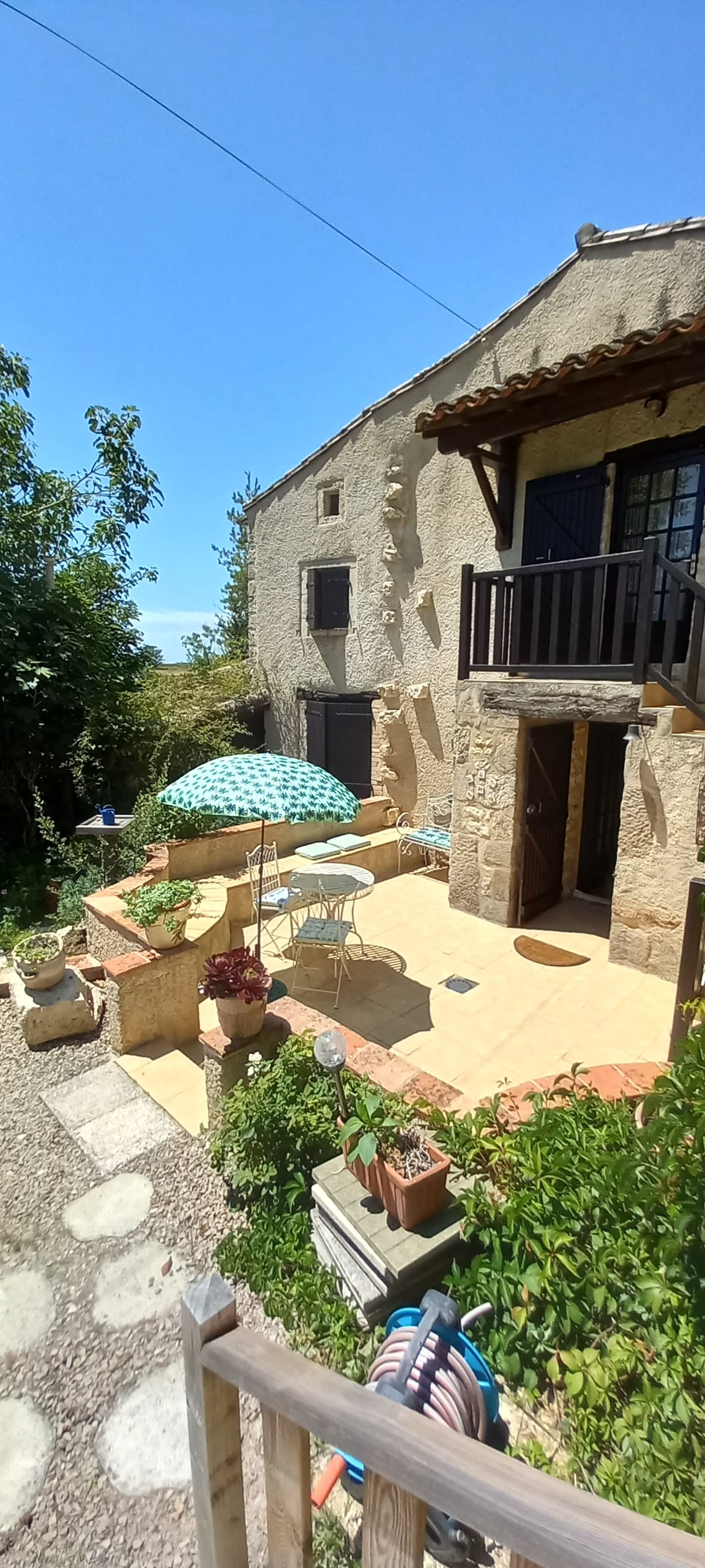 A charming exterior of a stone house is featured, showcasing a sunlit patio adorned with a blue and white patterned umbrella. A small table and chairs are visible, surrounded by lush greenery and flower pots, creating a serene outdoor space for relaxation.