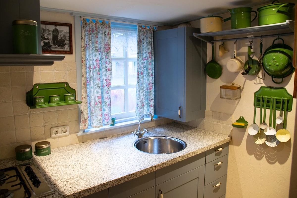 A functional kitchen is presented, featuring a granite countertop and stainless steel sink. Green cookware and utensils hang from wall hooks, with floral curtains framing a window allowing natural light. Shelves hold additional kitchen items, while the overall design combines practicality with a homey aesthetic.
